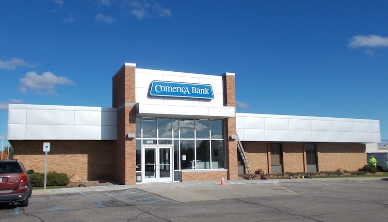 Front view of a Comerica Bank branch with a glass entry, brick walls, and a blue Comerica Bank sign against a clear blue sky. Parking lot in foreground.
