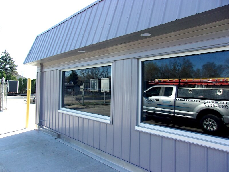Exterior of a light gray metal-clad building with two large windows; reflection shows a pickup truck with a ladder rack in the right window.